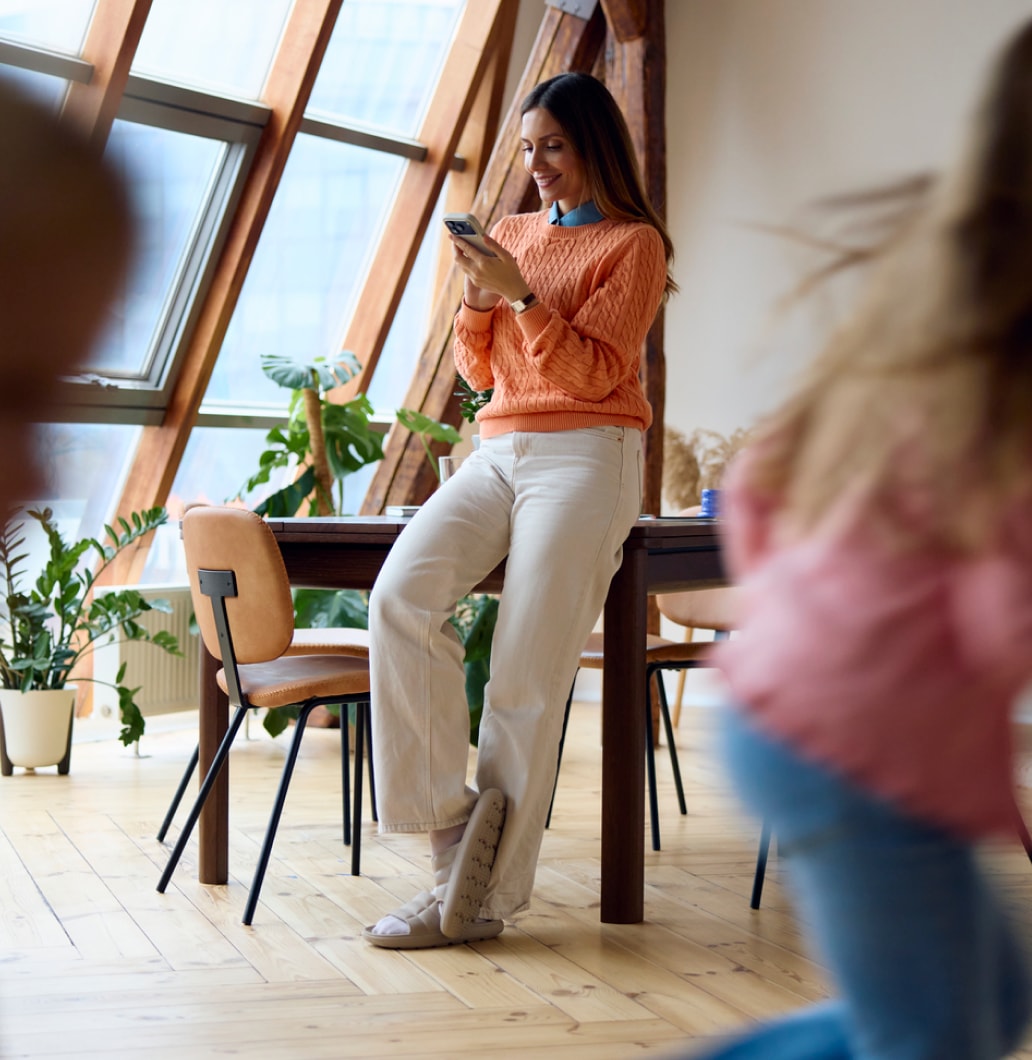 A woman checks her phone in a dining room while her children play